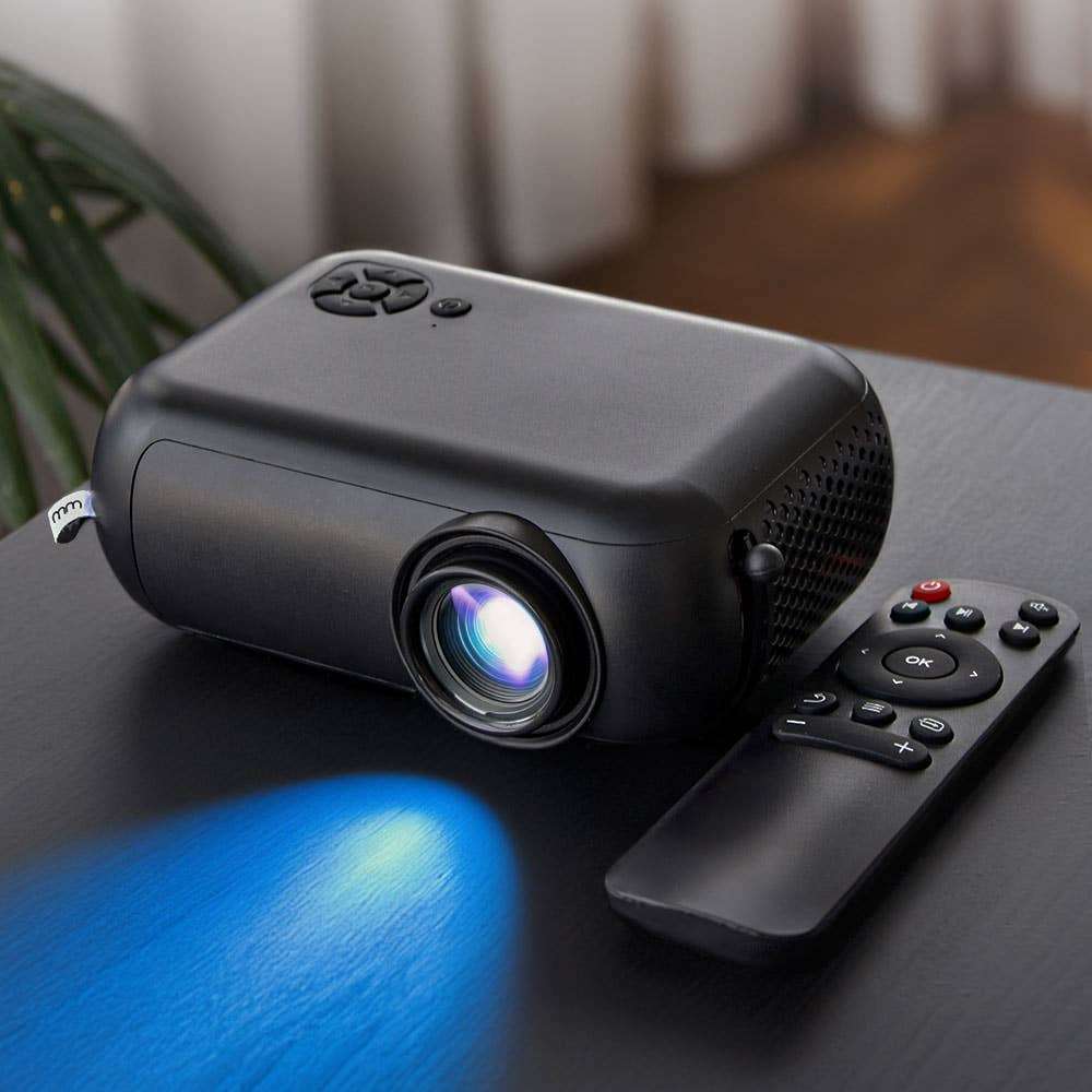 Black smart mini projector with illuminated lens and remote control on a dark tabletop.
