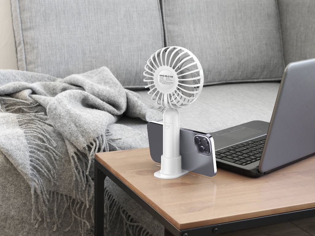 White portable fan on a wooden table next to a laptop and gray blanket on a couch.
